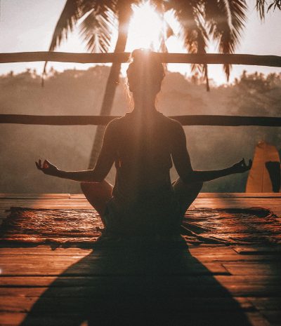 Eine Frau macht Yoga-Meditation bei Sonnenuntergang vor einer Palme.